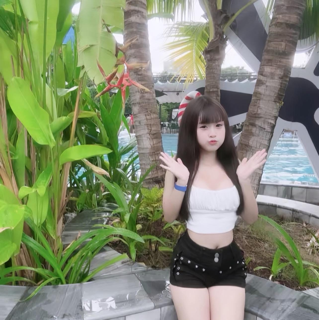 The work is an outdoor photo, the image of a girl wearing a white two-string shirt combined with black shorts, posing with her hands raised to the sides of her face with long dark brown hair, sitting next to the pool area. Behind the character is a row of